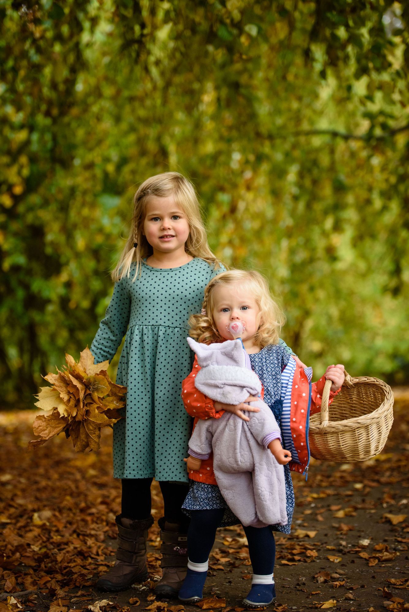 Zwei kleine Mädchen mit Herbstlaub, Korb und Puppe auf einem laubbedeckten Weg