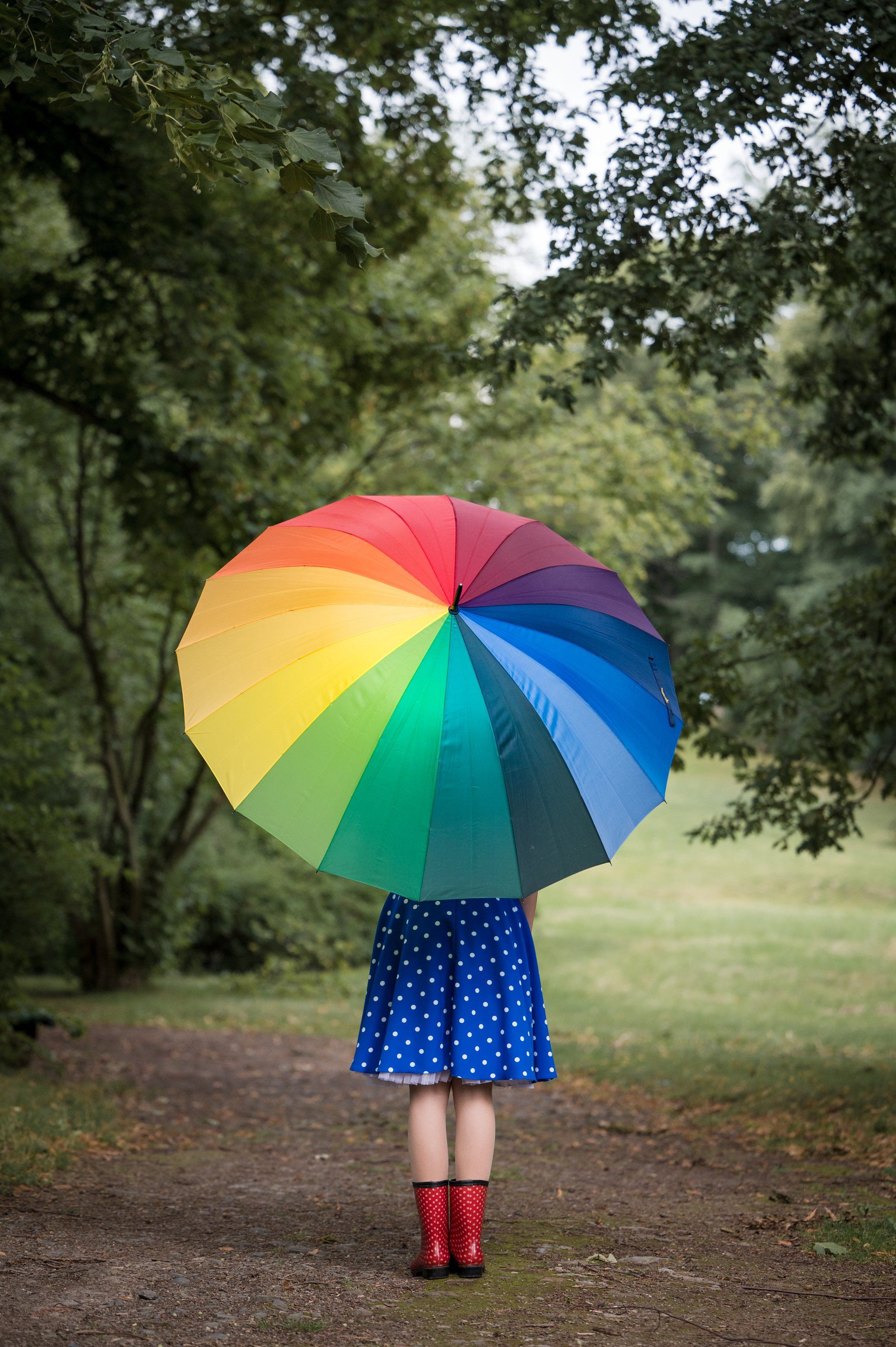 Mädchen mit Regenbogenschirm und roten Gummistiefeln von hinten im Park
