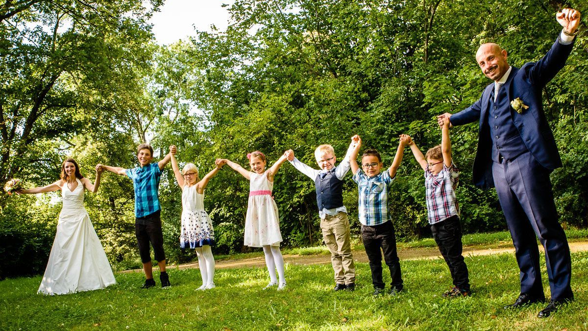 Braut und Bräutigam stehen mit sechs Kindern Hand in Hand in einer Reihe im Park
